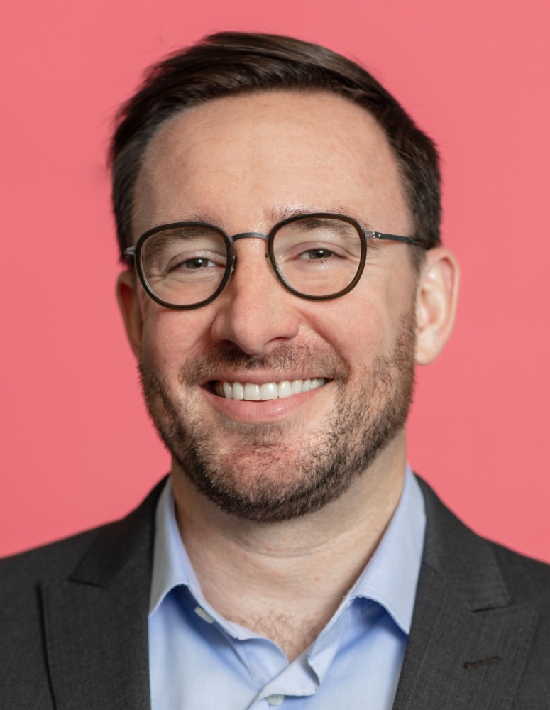 A person in a suit and glasses smiles against a pink background.